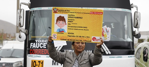 Persona con discapacidad severa sostiene pancarta de carné conadis amarillo frente a un bus de transporte público