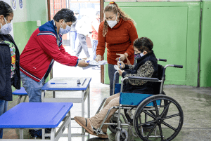 Una mujer adulta mayor con discapacidad física, usuaria de silla de ruedas, ejerce su derecho al sufragio con el apoyo de una persona miembro de mesa.