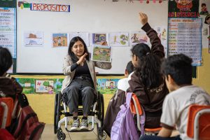 Una docente con discapacidad física que utiliza silla de ruedas se encuentra en un salón de clase frente a sus alumnos. Se encuentra narrando una historia mientras sostiene con la mano izquierda un dibujo que muestra a un personaje de la novela "El Hobbit".