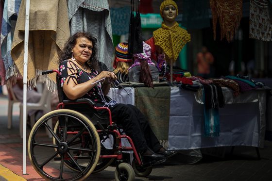 Una mujer con discapacidad usuaria de silla de ruedas se encuentra tejiendo a dos agujas sentada frente a su emprendimiento de productos textiles.