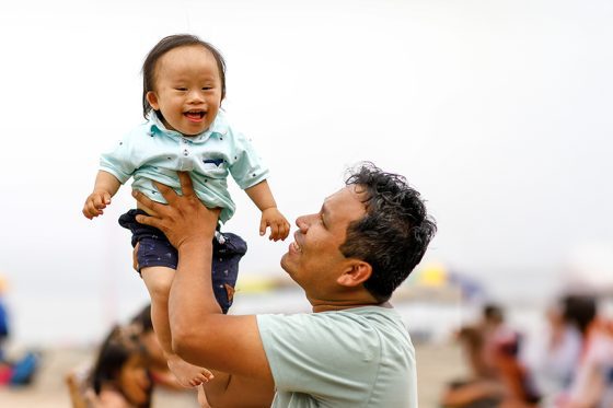 En la playa, un padre emocionado alza en brazos a su bebé. El bebé es un niño con síndrome de Down. Ambos están felices y sonrien.