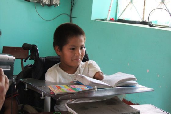 Un niño con discapacidad usuario de silla de ruedas revisa el contenido de su libreta de notas frente a su carpeta en el salón de clases.