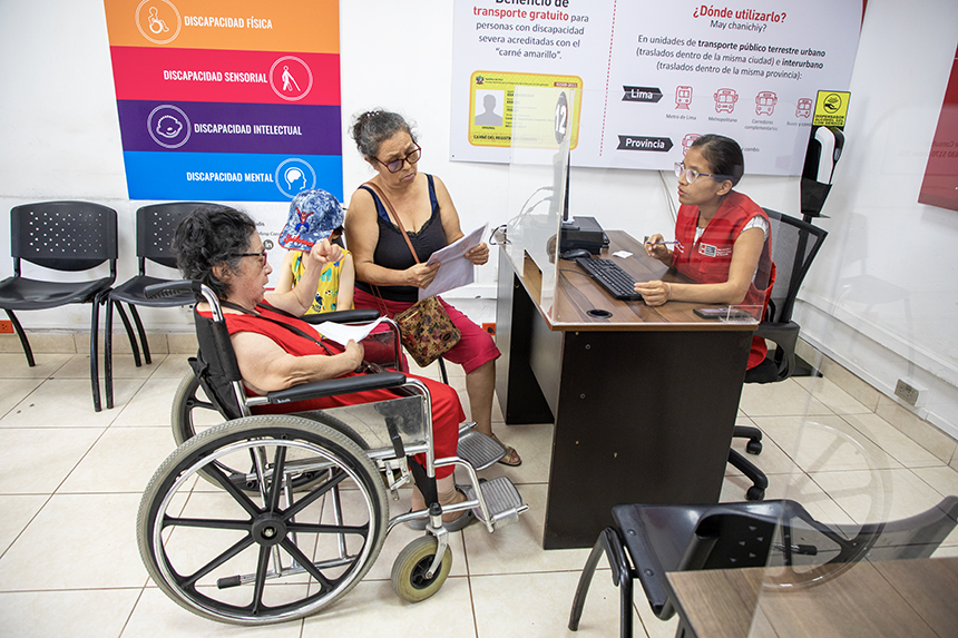 Un amujer adulta mayor con discapacidad física y que es usuaria de silla de ruedas, se encuentra acompañada de una familiar y escucha atentamente las indicaciones de una servidora pública del Conadis.