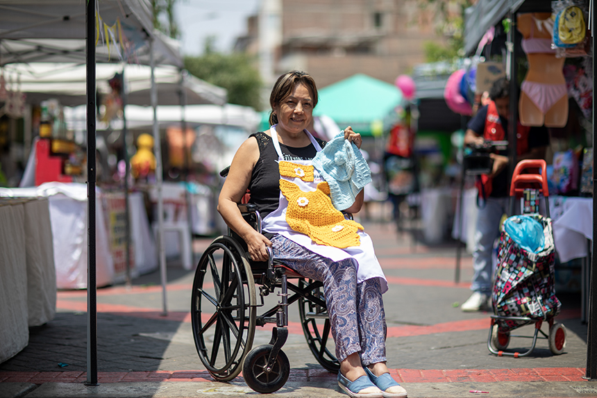 Mujer con discapacidad física, usuaria de silla de ruedas, presenta los productos de su emprendimiento en una feria.