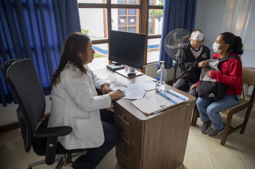 Una profesional de la salud atiende en su consultorio a un joven con mascarilla y a su acompañante sentados frente a su escritorio.