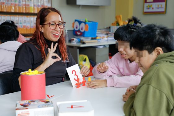 Maestra enseña a dos niños en una institución educativa. Con una mano sostiene una cartilla que tiene dibujado el número tres, mientras que con la otra muestra tres dedos, haciendo alusión a dicha cantidad.