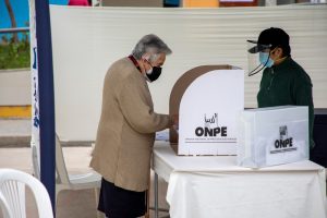 Una persona adulta mayor emite su voto en una mesa de sufragio de la ONPE, mientras otra persona con protector facial observa el proceso.