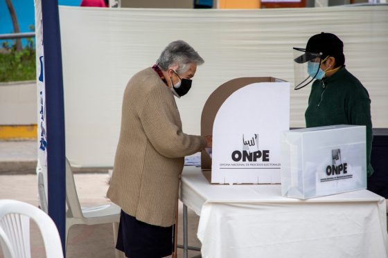 Una persona adulta mayor emite su voto en una mesa de sufragio de la ONPE, mientras otra persona con protector facial observa el proceso.