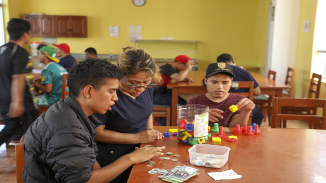 Una educadora ayuda a un joven a contar monedas y billetes en una mesa, mientras otro estudiante manipula piezas de plástico de colores en un aula.