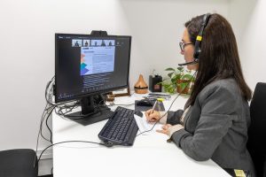 Una mujer participa en una videoconferencia desde su oficina, usando audífonos con micrófono y tomando notas mientras observa una presentación en su monitor. En el escritorio tiene su teclado, documentos y algunos objetos de oficina.