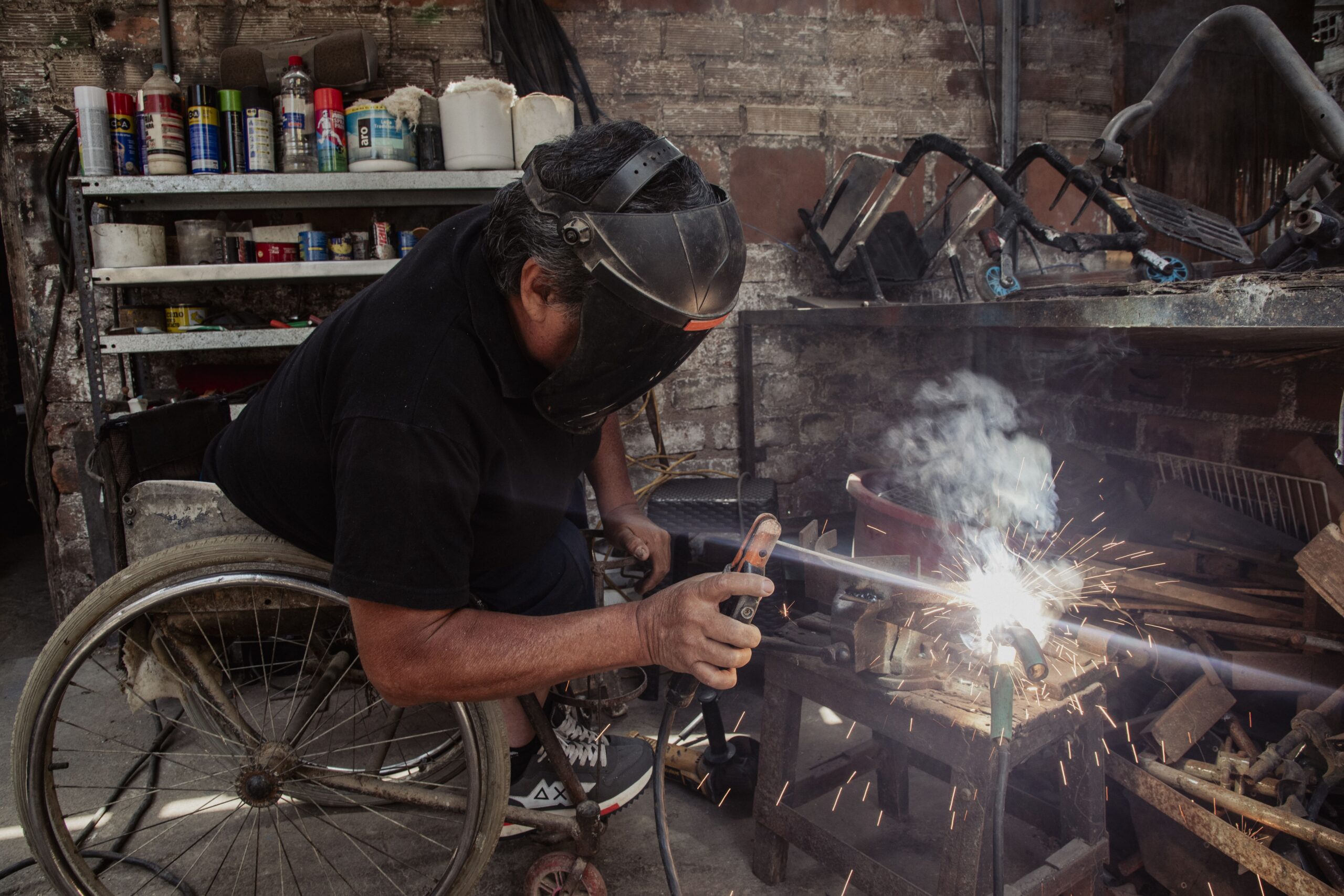 Un soldador en silla de ruedas, usando una careta protectora levantada hacia arriba, realiza trabajos de soldadura generando chispas y humo en su taller.