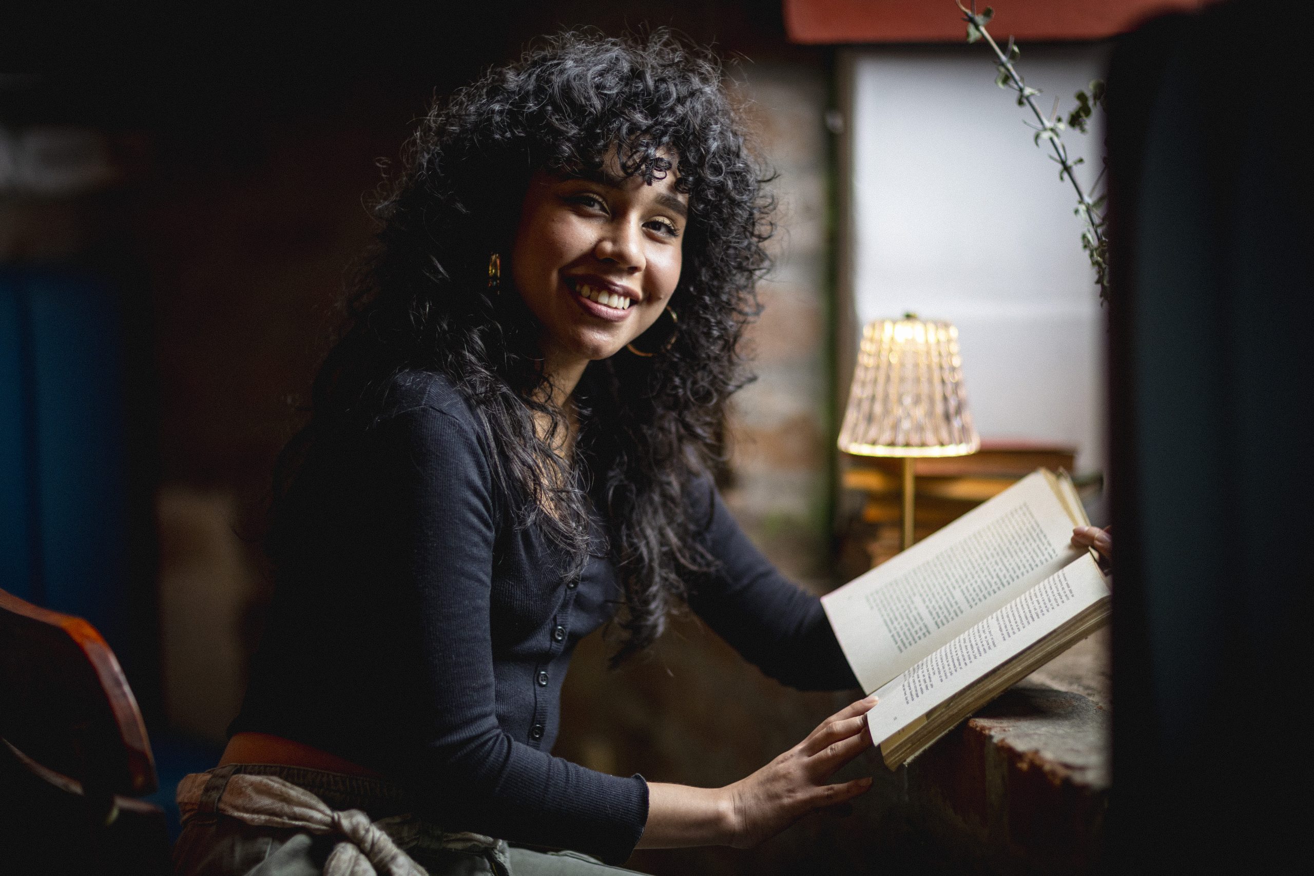 La imagen muestra a una mujer joven leyendo un libro en un ambiente cálido y acogedor. Está sentada junto a una ventana de luz tenue y una pequeña lámpara que ilumina suavemente la escena