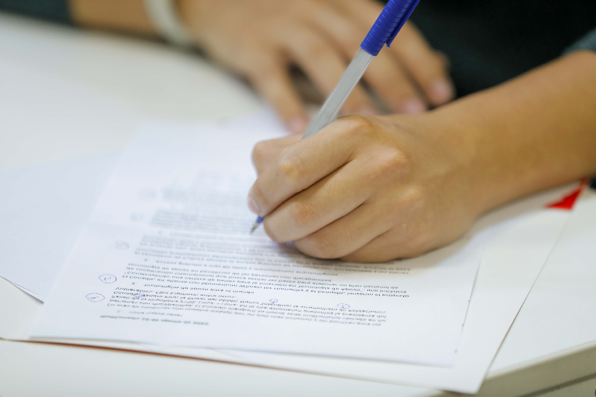 Mano de una persona escribiendo con un bolígrafo azul sobre un documento impreso con anotaciones y marcas, sobre una mesa blanca.