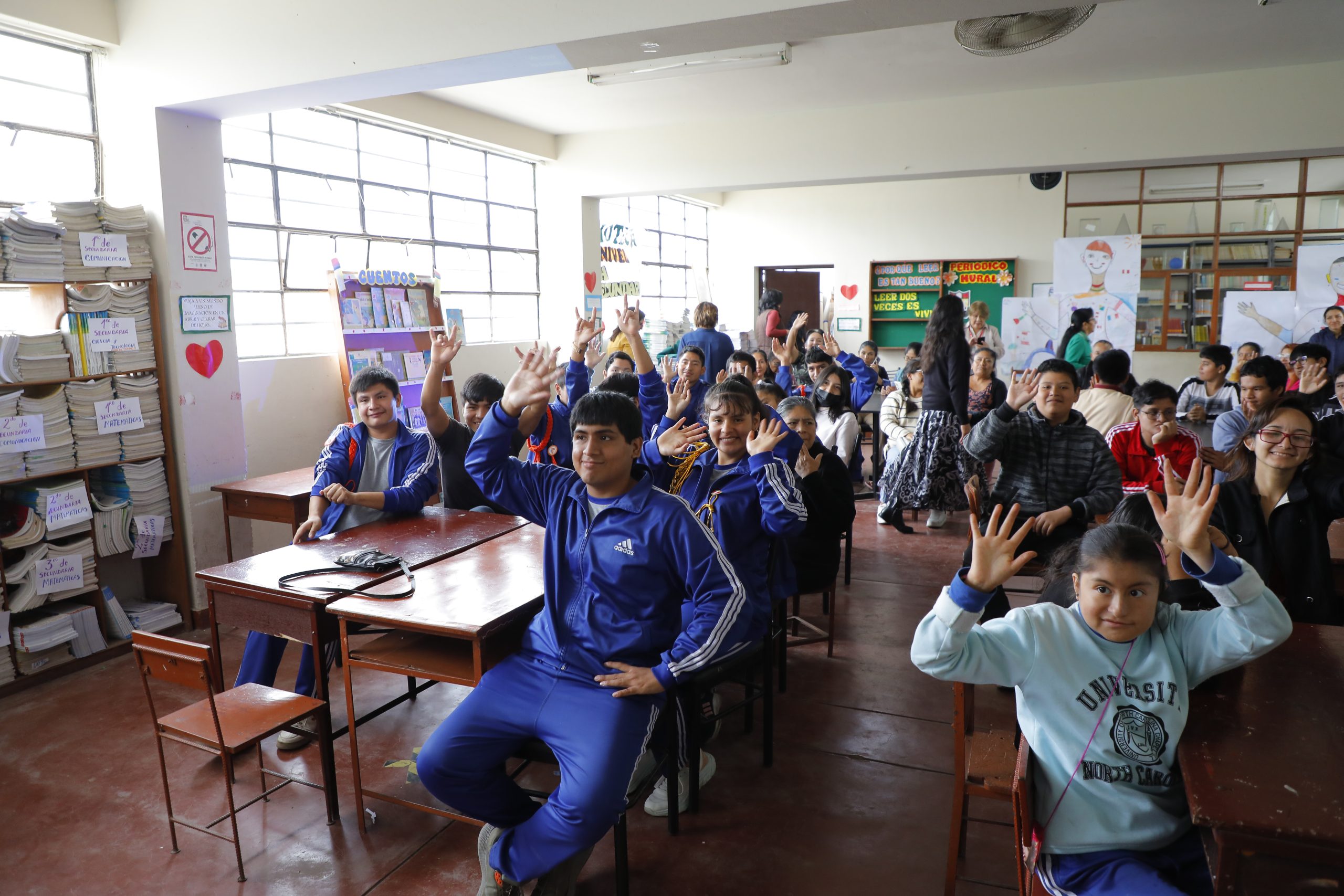 Estudiantes sentados en un aula escolar levantan la mano y sonríen durante una actividad educativa grupal.