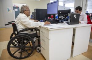 Un adulto mayor en silla de ruedas se encuentra trabajando en una computadora.