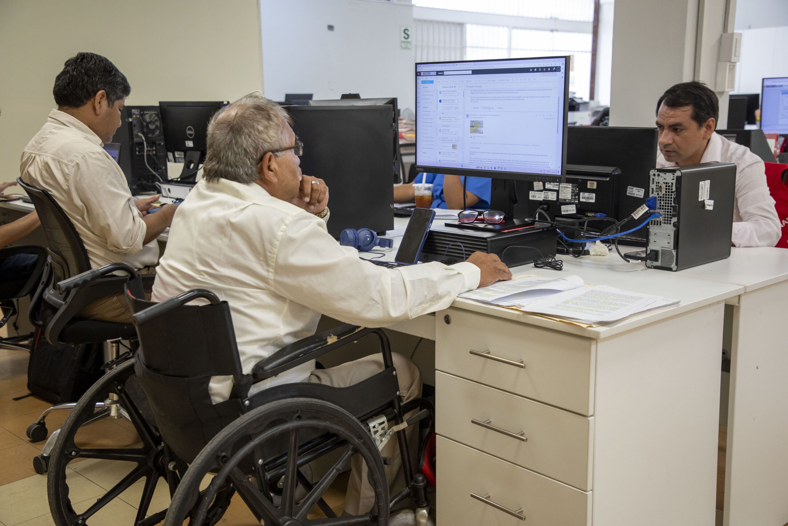Tres hombres trabajando en un entorno de oficina con computadoras; en primer plano, un hombre en silla de ruedas observa la pantalla de un monitor.