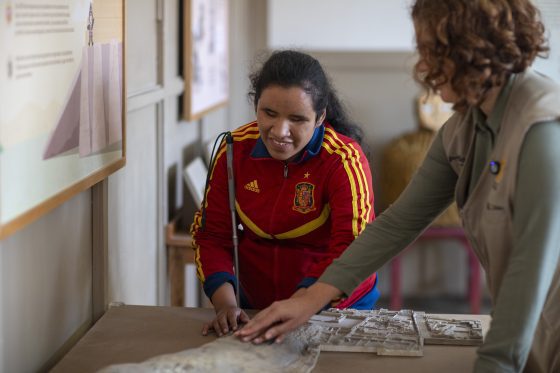 Mujer con discapacidad visual, usando bastón, toca una maqueta en relieve mientras otra mujer la guía con la mano.