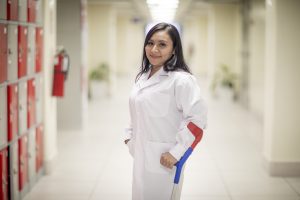 Mujer con bata médica en un pasillo de hospital, utilizando una muleta en el brazo derecho y sonriendo a la cámara.