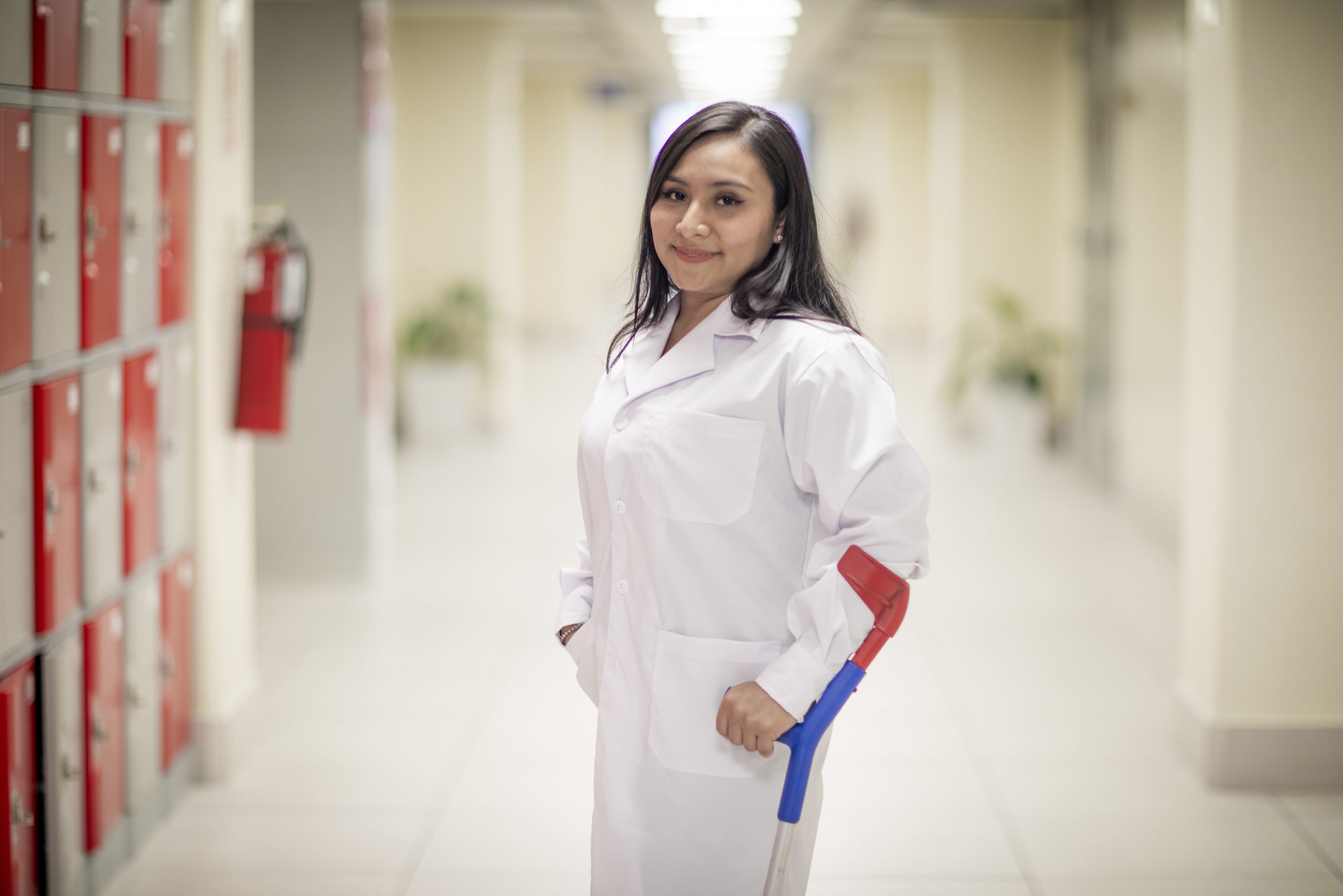 Mujer con bata médica en un pasillo de hospital, utilizando una muleta en el brazo derecho y sonriendo a la cámara.