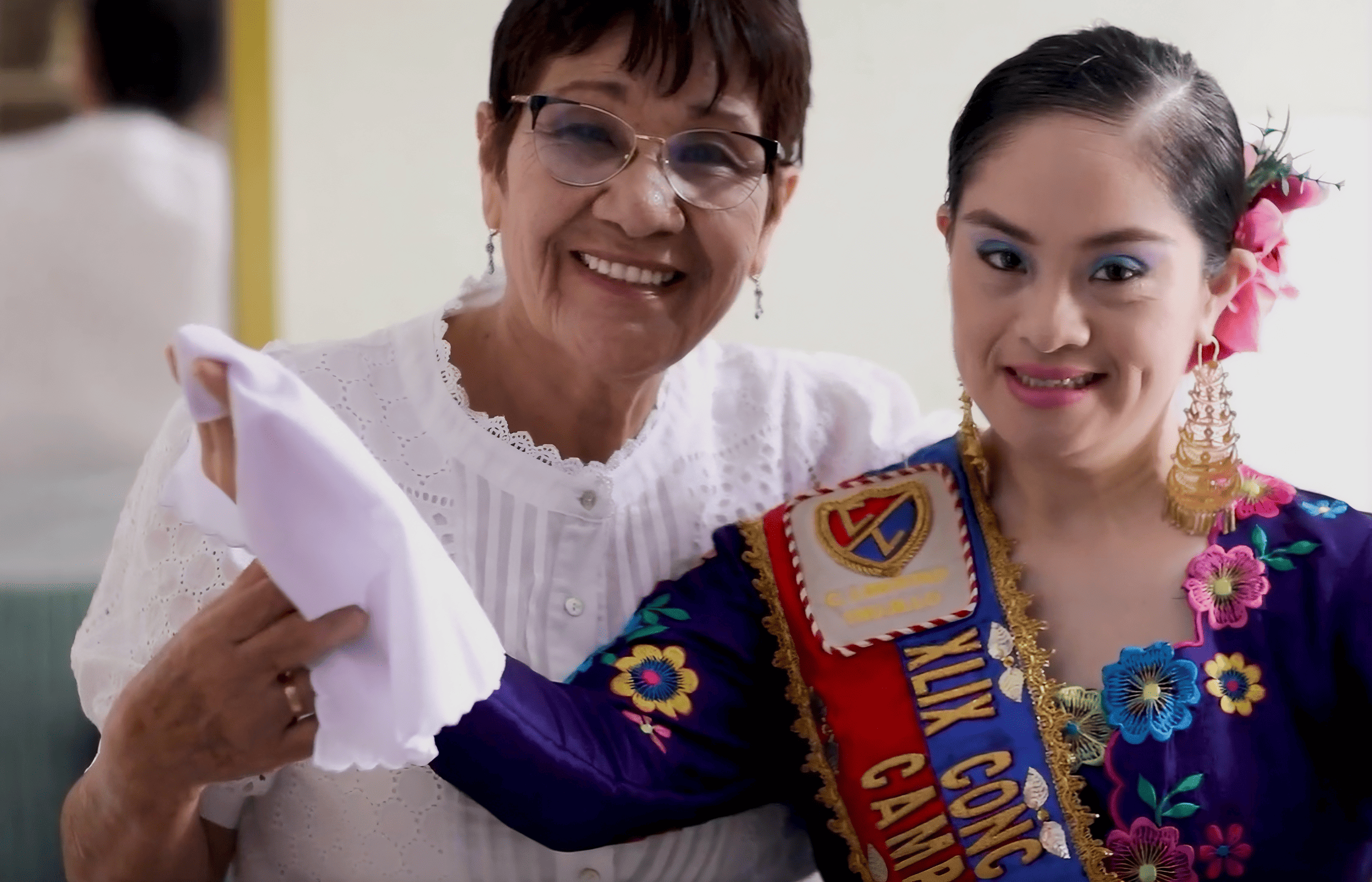Dos mujeres sonríen juntas frente a la cámara. A la izquierda, una mujer mayor con gafas y blusa blanca sostiene un pañuelo. A la derecha, una mujer joven vestida con un traje tradicional colorido, con flores bordadas y un adorno en el cabello, lleva una banda que cruza su pecho. Ambas se ven felices y cercanas, como si celebraran un evento cultural o reconocimiento.