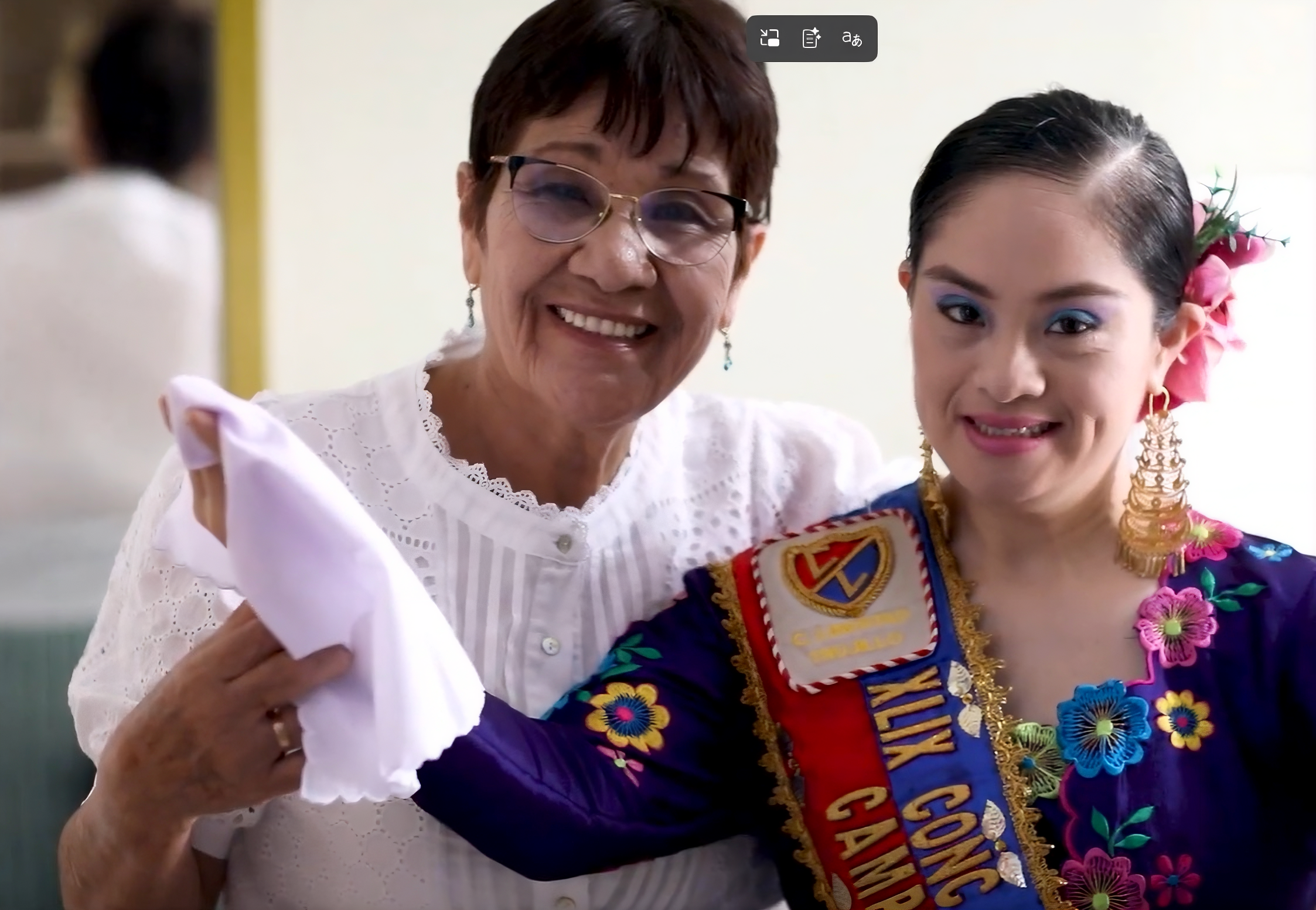 Dos mujeres sonríen juntas frente a la cámara. A la izquierda, una mujer mayor con gafas y blusa blanca sostiene un pañuelo. A la derecha, una mujer joven vestida con un traje tradicional colorido, con flores bordadas y un adorno en el cabello, lleva una banda que cruza su pecho. Ambas se ven felices y cercanas, como si celebraran un evento cultural o reconocimiento.