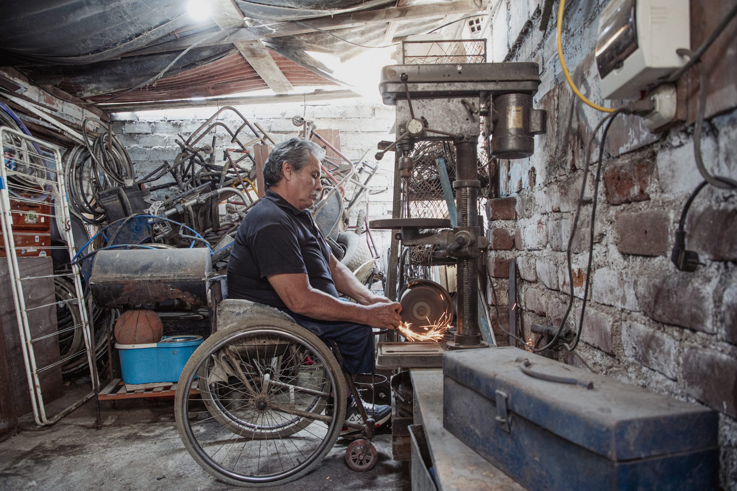 Hombre en silla de ruedas trabajando con una máquina de esmeril en un pequeño taller lleno de herramientas, piezas metálicas y bicicletas, mientras las chispas saltan al afilar o cortar un objeto.
