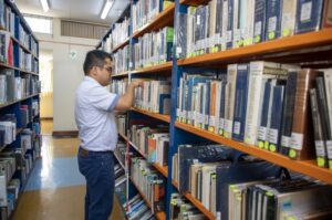 Una persona busca un libro en los estantes de una biblioteca, rodeada de estanterías con libros.