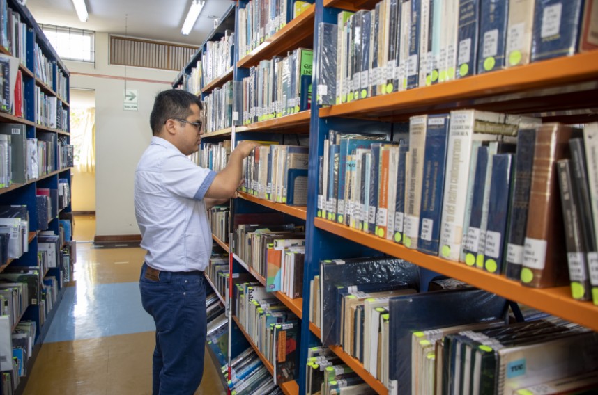 Una persona busca un libro en los estantes de una biblioteca, rodeada de estanterías con libros.
