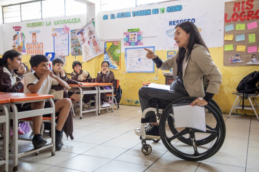 Una profesora en silla de ruedas enseña a un grupo de niños y niñas atentos en un aula escolar decorada con afiches educativos.