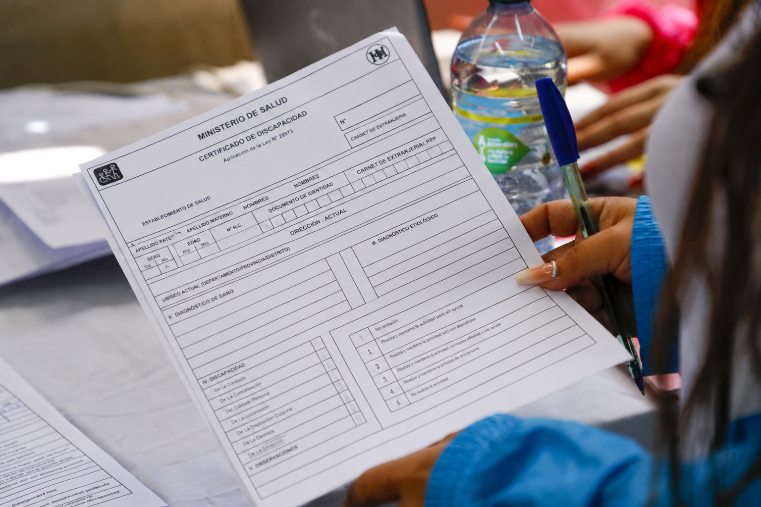 Una persona sostiene un formulario en blanco del Ministerio de Salud titulado 'Certificado de Discapacidad', junto a una lapicero azul y una botella de agua sobre una mesa.