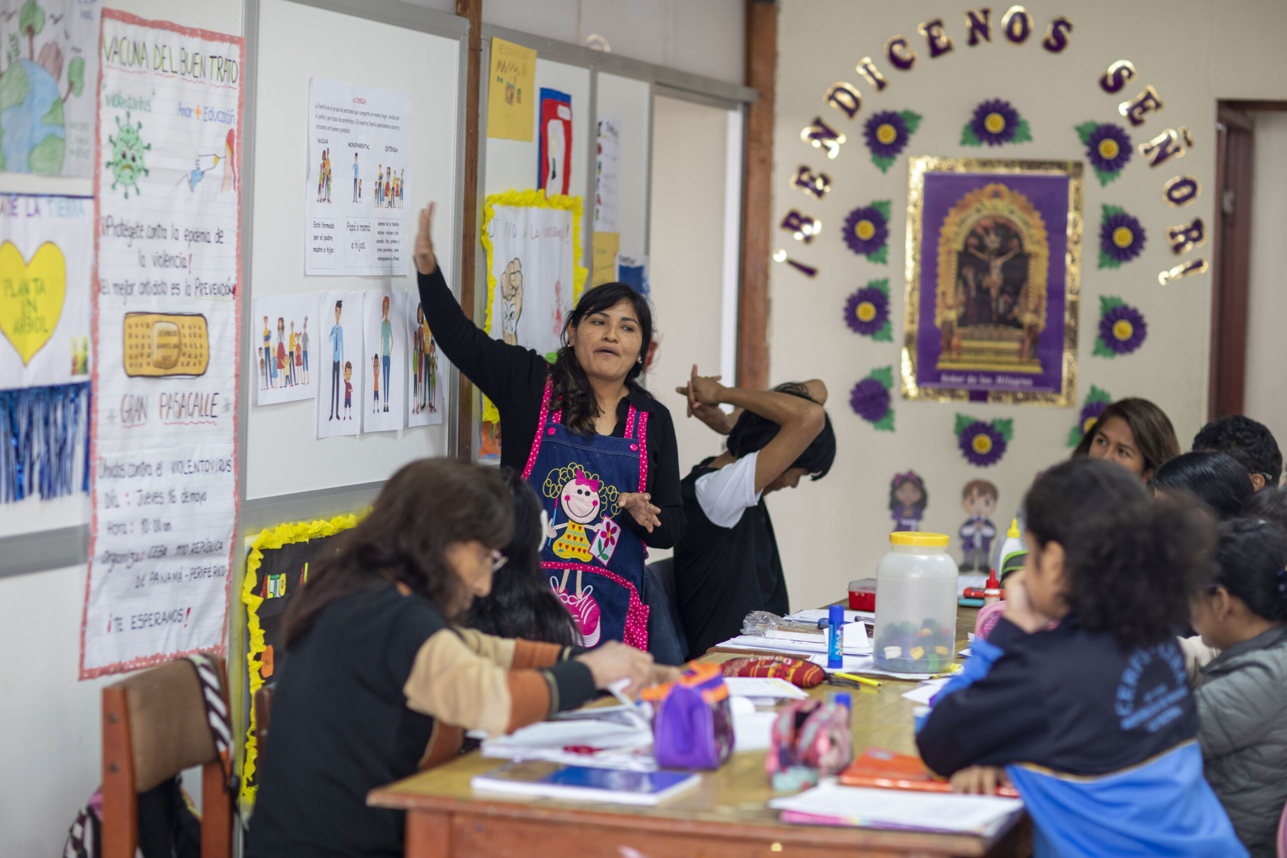 Una profesora con delantal le enseña a un grupo de estudiantes sentados alrededor de una mesa en un aula decorada con afiches educativos y religiosos.