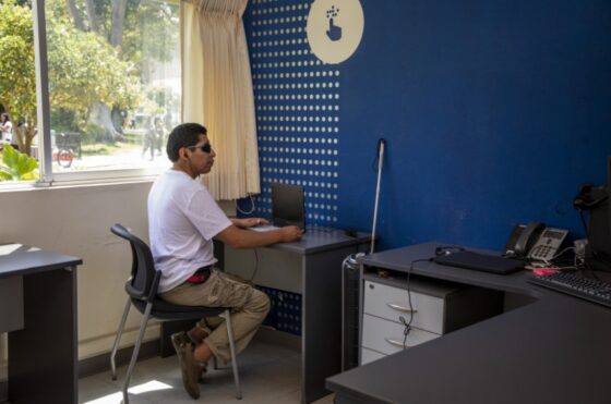 La imagen muestra a una persona con discapacidad visual frente a una computadora portatil.