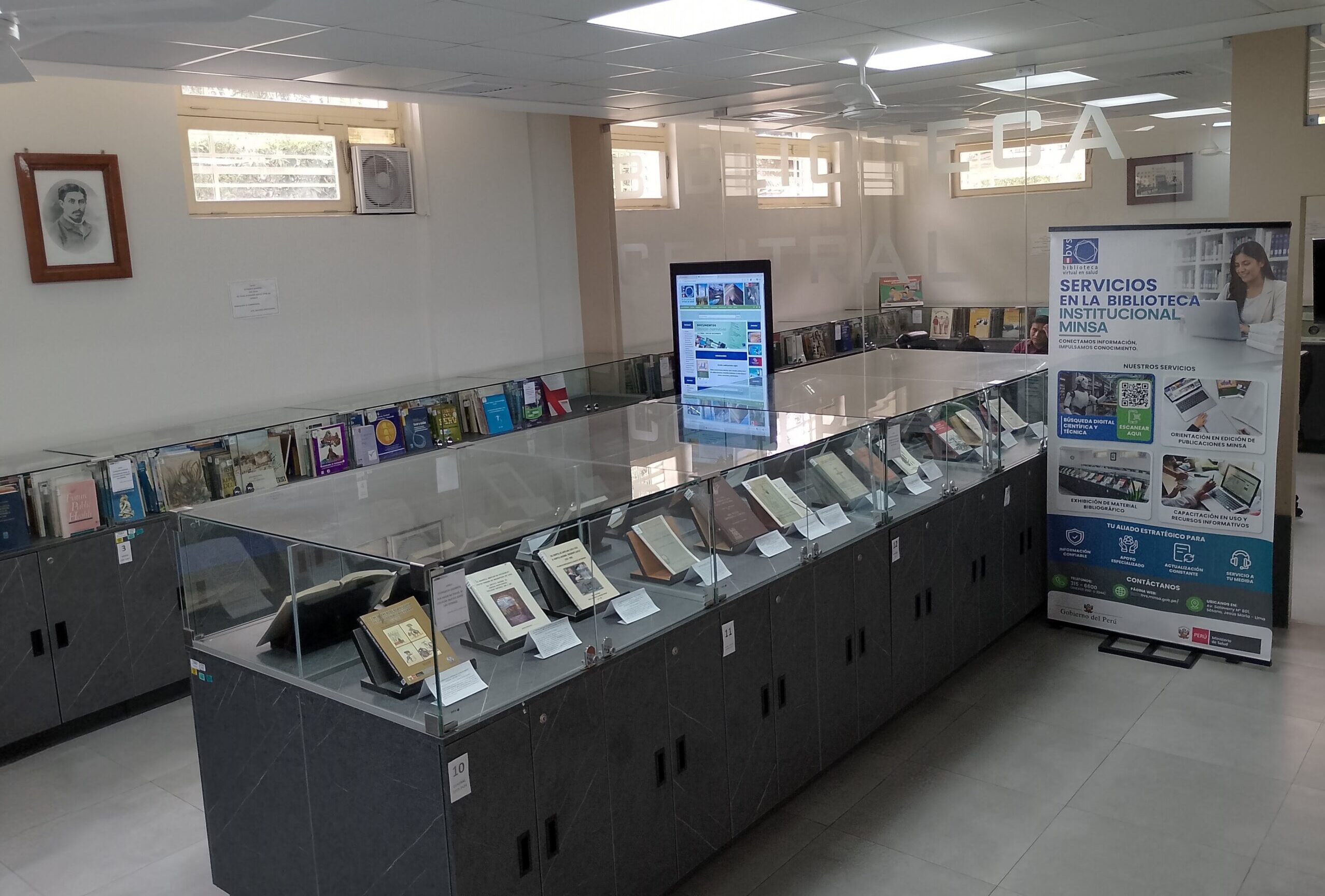 Vista interior de la Biblioteca Institucional del Ministerio de Salud del Perú (MINSA), mostrando vitrinas de vidrio gris con libros expuestos, estantes de lectura al fondo y un banner informativo.