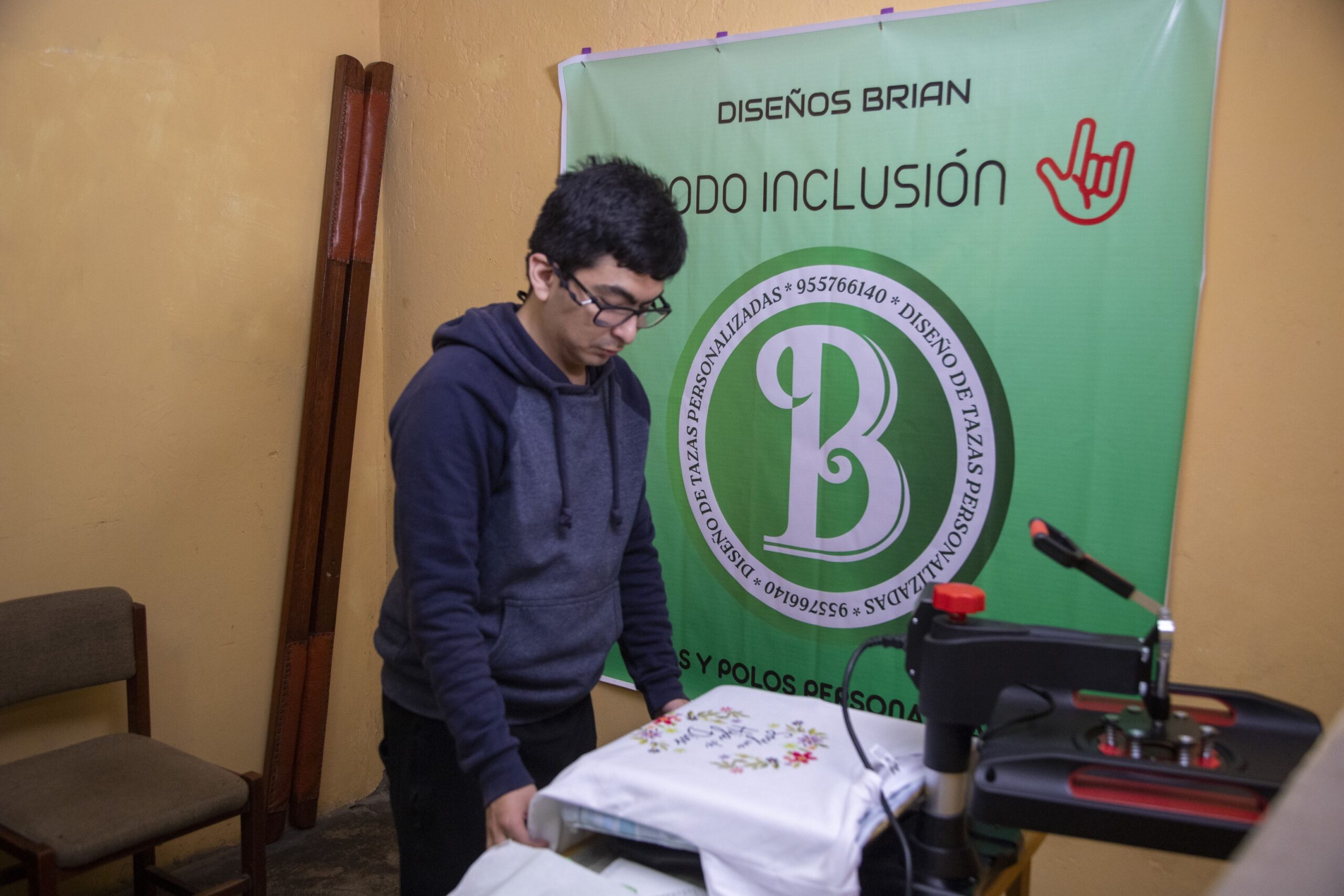 Un joven con lentes acomoda una prenda blanca sobre una estampadora térmica frente a un banner publicitario de su emprendimiento «Diseños Brian - Modo Inclusión».