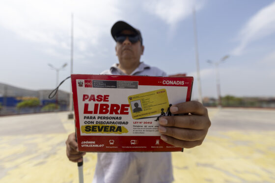 Un hombre con gafas de sol sostiene hacia la cámara un cartel del CONADIS que anuncia el "Pase Libre para personas con discapacidad severa".