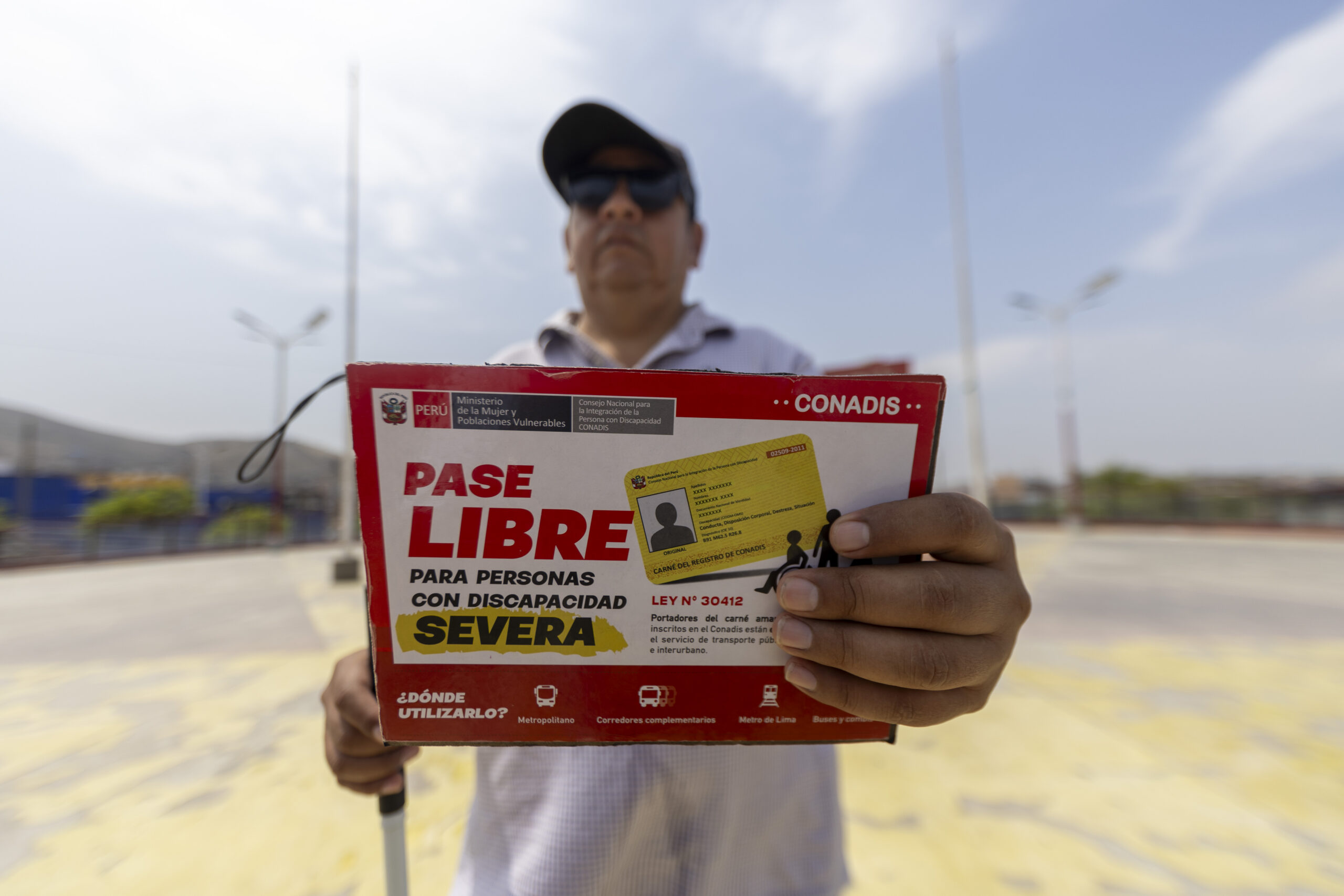 Un hombre con gafas de sol sostiene hacia la cámara un cartel del CONADIS que anuncia el "Pase Libre para personas con discapacidad severa".