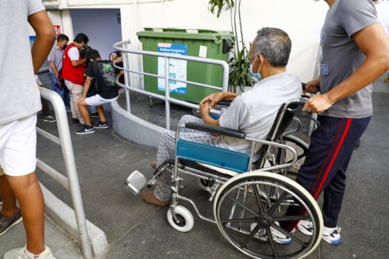 Un adulto mayor en silla de ruedas es trasladado por una persona por una rampa de accesibilidad en un espacio público.