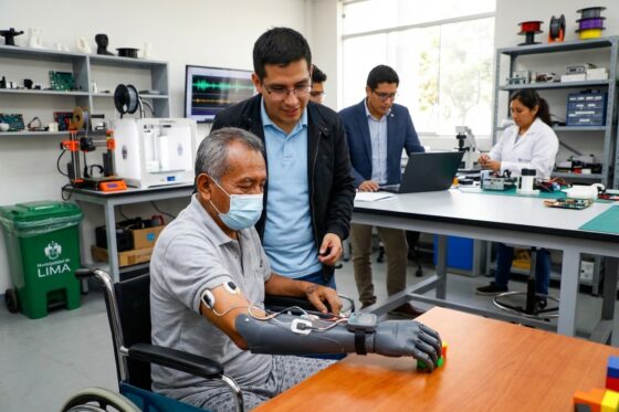 Un paciente en silla de ruedas y con mascarilla prueba una mano protésica impresa en 3D en un laboratorio. Un investigador lo guía